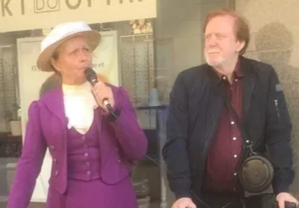 En kvinna iklädd en lila dräkt och en vit hatt hon håller i en mikrofon, en man i brunt hår och skägg iklädd en mörk jacka och en vinröd skjorta.
