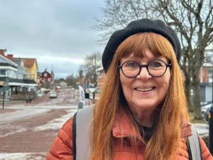 En kvinna i långt rödlätt hår och glasögon, hon har en svart basker på huvudet, stadsmiljö i bakgrunden.