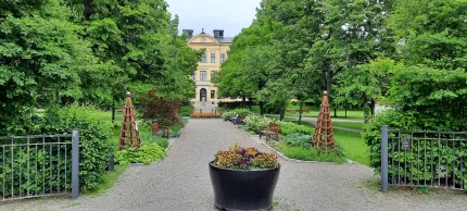 Entrén till en park, järnstaket, en grusgång med planteringar på bägge sidor, i slutet av gången en gul stenbyggnad.