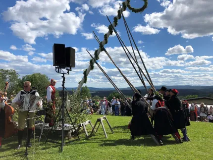 Majstången reses av män och kvinnor klädda i folkdräkt, besökare i bakgrunden, bånande berg och en blå himmel med några vita moln.