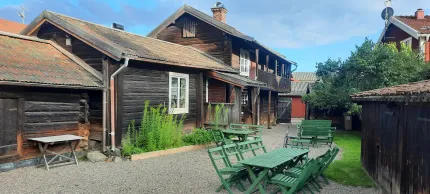 Gråbruna timmerbyggnader på en gård, gröna trädgårdsmöbler i trä i framkant.