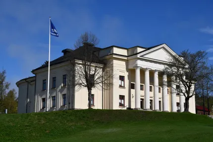 Vit klassisk byggnad med hög pelarentré och flaggstång på grön gräsmatta under klarblå himmel..
