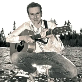 Person på huk vid en stenig strand håller en akustisk gitarr; trädlinje och vatten syns i bakgrunden..
