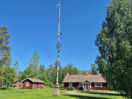 En hög majstång på en gräsmatta framför röda gårdsbyggnader, omgiven av träd under klarblå himmel..