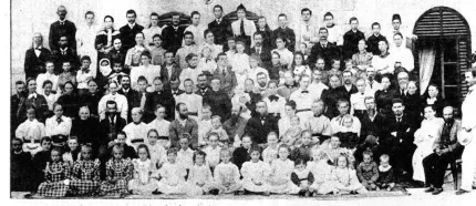 Svartvitt gruppfoto med många människor i flera rader utanför en byggnad med fönsterluckor och dörr..