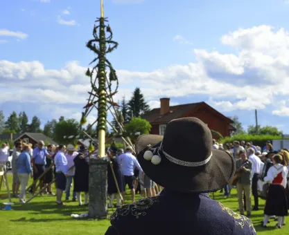 En person i svart hatt i förgrunden ser en majstång och en folksamling på en gräsmatta under blå himmel..