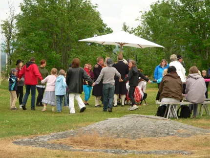 En grupp människor håller varandra i händerna i en ring på en gräsmatta, med stort parasoll och träd i bakgrunden..