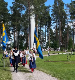 Folk i folkdräkter går i procession med svenska flaggor på en grusväg genom en grön park med tallskog..