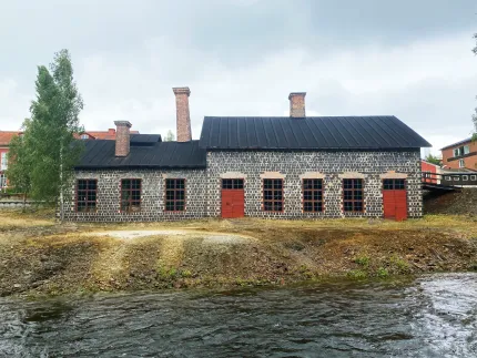 Lång stenbyggnad med svart plåttak, röda dörrar och fönster vid en strömmande å under grå himmel..