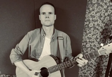 En person sitter och spelar akustisk gitarr med axelrem mot mörk bakgrund och blommigt mönster till höger..