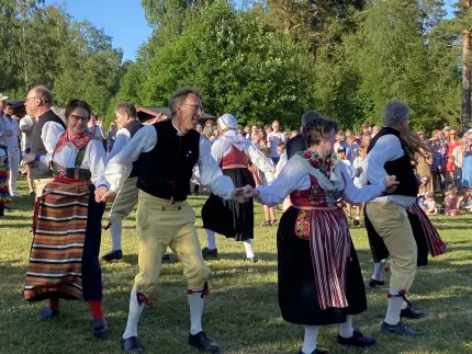 Grupp människor i traditionella folkdräkter dansar hand i hand på en gräsmatta, med publik och träd i bakgrunden..