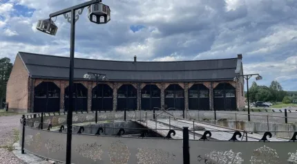 Stor tegelbyggnad med svarta portar bakom en rund fontän med gångbro och lyktstolpar under molnig himmel..