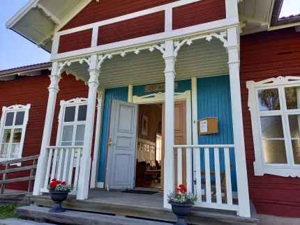 Entréveranda till ett rött trähus med vit snickarglädje, blå panel, öppen dörr, brevlåda och blomkrukor..