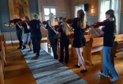 En grupp ungdomar spelar fiol i en rad inne i en kyrka, med träbänkar, fönsterljus och målning på väggen..