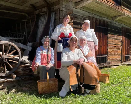 Fem personer i traditionella kläder sitter och står vid en gammal timmerstuga med vagnshjul och korgar i sol..