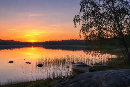 Solnedgång över en spegelblank sjö med vass och stenar i vattnet, samt en roddbåt vid strandkanten..