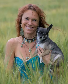En kvinna i turkos klänning och smycken håller en liten hund i ett fält med högt grönt gräs..