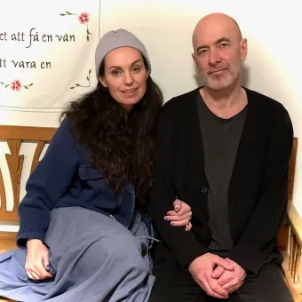 Två personer sitter tätt ihop på en träbänk inomhus, med varma kläder och en väggbonad i bakgrunden..