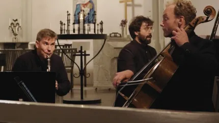 Tre musiker spelar klarinett och cello i en kyrkinteriör med altare, ljusstakar och kors i bakgrunden..