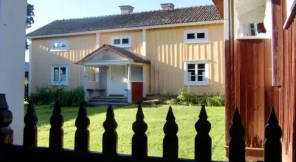 Gul träbyggnad med vita fönster och liten farstukvist, sedd över en gräsmatta bakom ett svart staket..