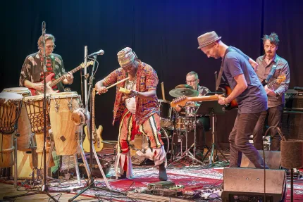 Fem musiker uppträder på en scen med trummor, slagverk och elgitarrer under färgade strålkastare..