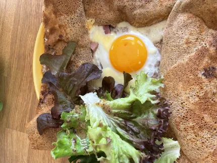 Närbild av en bovetegalett med stekt ägg, smält ost och skinkbitar, serverad med blandad sallad på tallrik..
