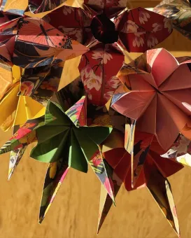 Närbild av flera origamiblommor i mönstrat papper med varma färger, upphängda mot en gul bakgrund..