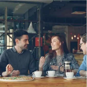 Tre personer sitter vid ett cafébord med kaffekoppar, en presskanna och en pizza medan de pratar och ler..