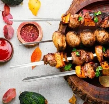 Närbild uppifrån av grillspett med kött, potatis och grönsaker på träbricka, med kryddor och sås bredvid..
