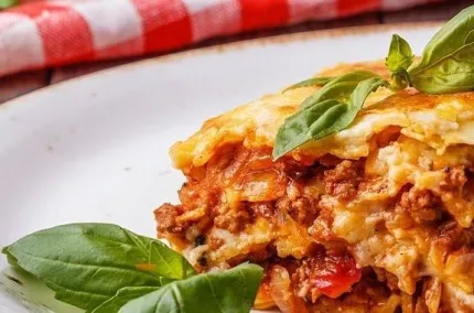 Närbild av en portion lasagne med köttfärssås och osttäcke, toppad med färsk basilika på vit tallrik..