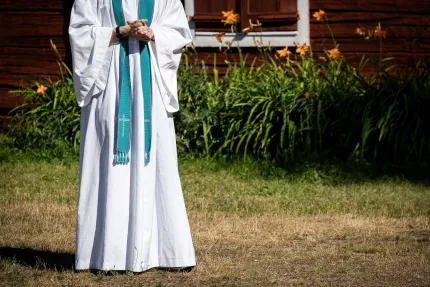 En person i vit prästkåpa med turkos stola står på gräs framför en träbyggnad och blommor..