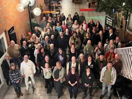 Stort gruppfoto med många personer som står i en inomhusgalleria med tegelvägg, lampor, trappa och växter..