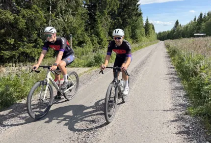 Två cyklister med hjälm och solglasögon cyklar på en solig grusväg genom skog och ängar..