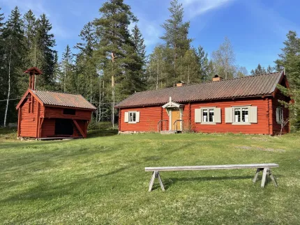 Två röda träbyggnader med tegelröda tak på en gräsmatta, omgivna av tallskog under klarblå himmel..