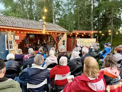Publik sitter på stolar framför en liten scen vid en röd lada med ljusslingor, omgiven av skog i skymning..