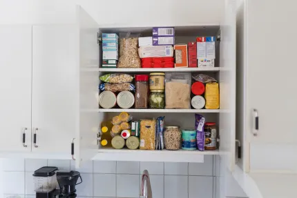 Öppet vitt köksskåp med tre hyllor fyllda med torrvaror, burkar, kartonger och påsar ovanför en diskbänk..