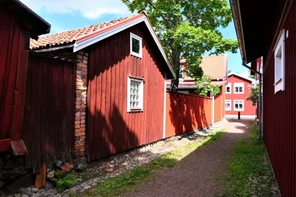 Smalt gångstråk mellan röda träbyggnader med tegeltak, ett träd som skuggar och blå himmel i bakgrunden..