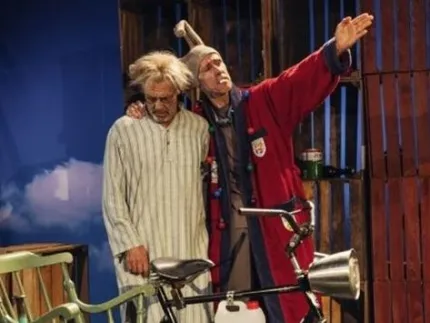 Två män står på en teaterscen vid en cykel med stol; en man i röd rock gestikulerar mot sidan..