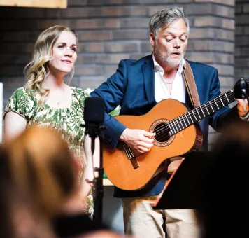 En man i kavaj spelar akustisk gitarr bredvid en kvinna i grön klänning, med mikrofon framför..
