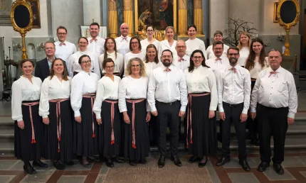 Grupporträtt av en kör i vit klädsel på trappsteg inne i en kyrka, med altartavla och pelare i bakgrunden..