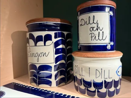 Tre blåvita keramiska burkar med trälock står på en hylla, med texten Lingon, Dill och Pill samt Sill i Dill..