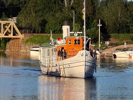 En vit ångbåt med orange styrhytt kör på en stilla kanal, med brygga, båtar och grön skog i bakgrunden..