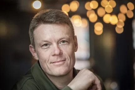 Närbildsporträtt av en man med kort hår och grön jacka, med varma suddiga ljuspunkter i bakgrunden..