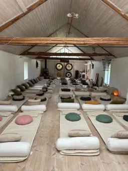 Långt rum med trägolv och synliga takbjälkar, yogamattor och kuddar i rader samt gongar längst fram..
