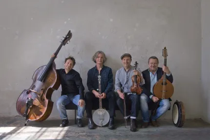 Fyra sittande musiker mot en sliten vägg med kontrabas, banjo, fiol, mandolin och en trumma på golvet..