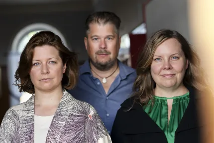 Porträtt av två kvinnor i förgrunden och en man bakom, stående i en korridor med mjukt ljus..