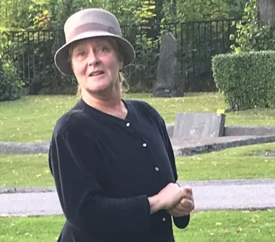 En kvinna i mörk kofta och beige hatt står på en gräsmatta med gravstenar och träd i bakgrunden..