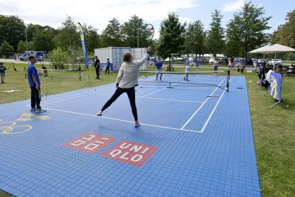En person slår ett slag på en blå temporär tennisbana i en park, med nät, bollar och åskådare runtom..