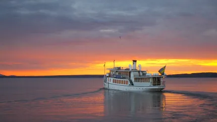 En vit passagerarbåt kör på stilla sjö i solnedgång, med orange himmel, moln och svag horisont..