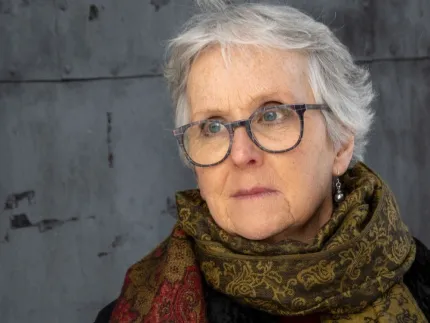 Närbild på en äldre kvinna med kort grått hår och runda glasögon, med mönstrad sjal mot grå vägg..